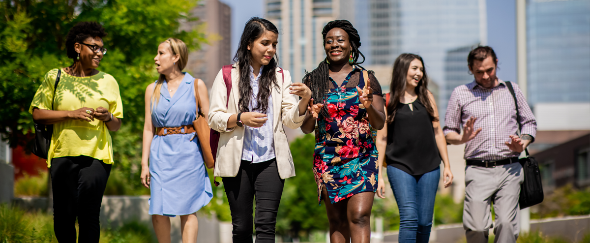 Image of 6 students walking toward the camera, chatting with each other, on a bright, sunny day outside on Auraria Campus.